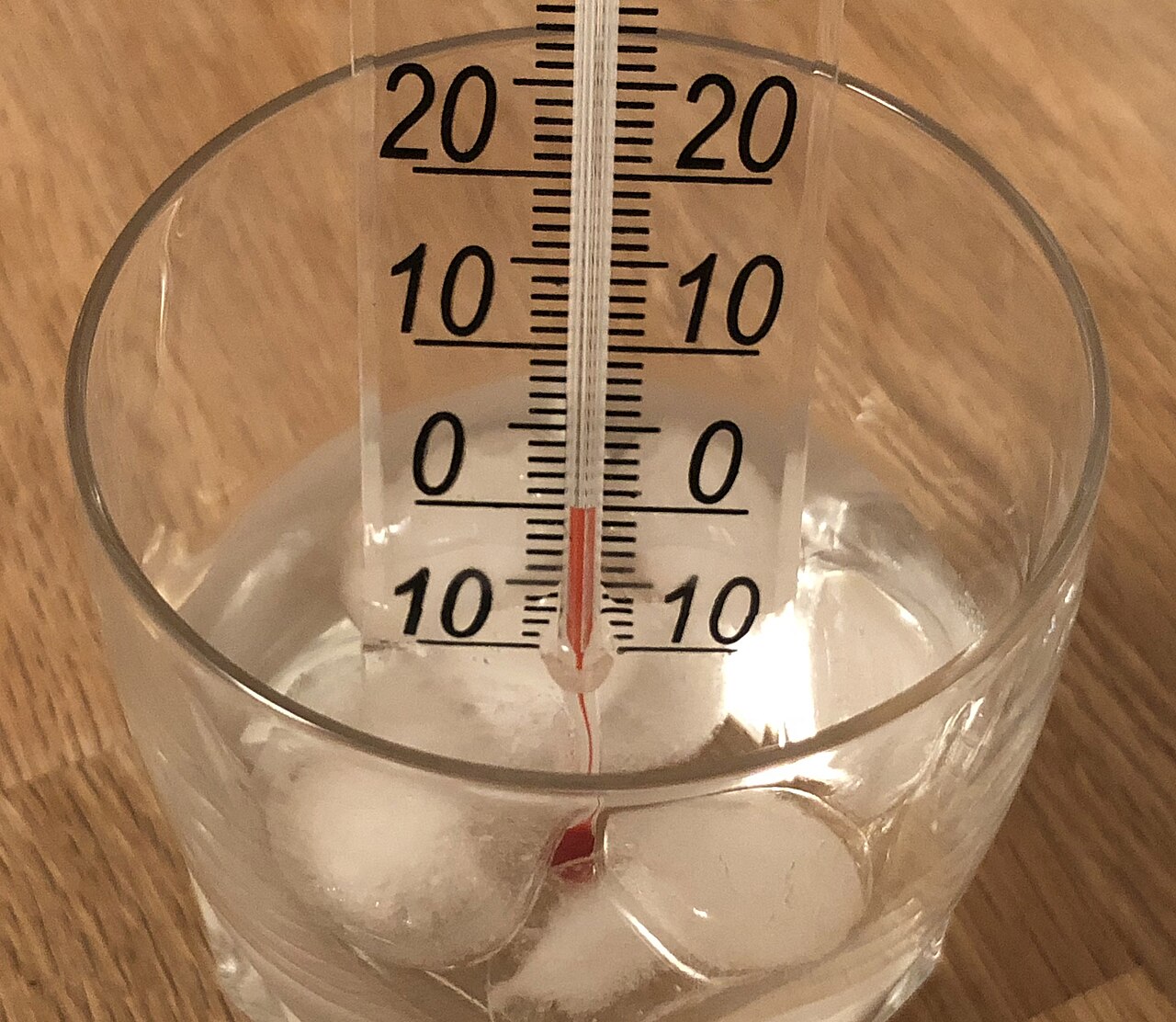 Glazen vloeistofthermometer in een glas met water en ijsblokjes, met een schaal van min tien tot twintig graden.